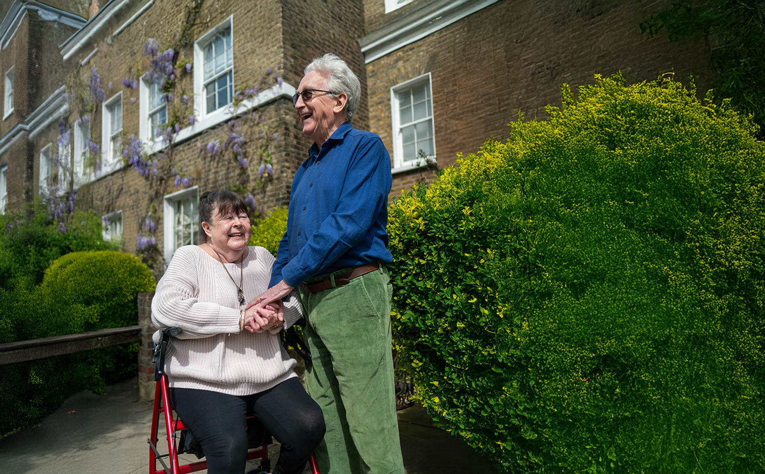 A documentary-style photograph of two elderly people interacting outdoors. A man in a blue shirt stands holding hands with a seated woman. They appear to be siblings sharing a moment of joy and relief, reflecting the temporary housing case story.