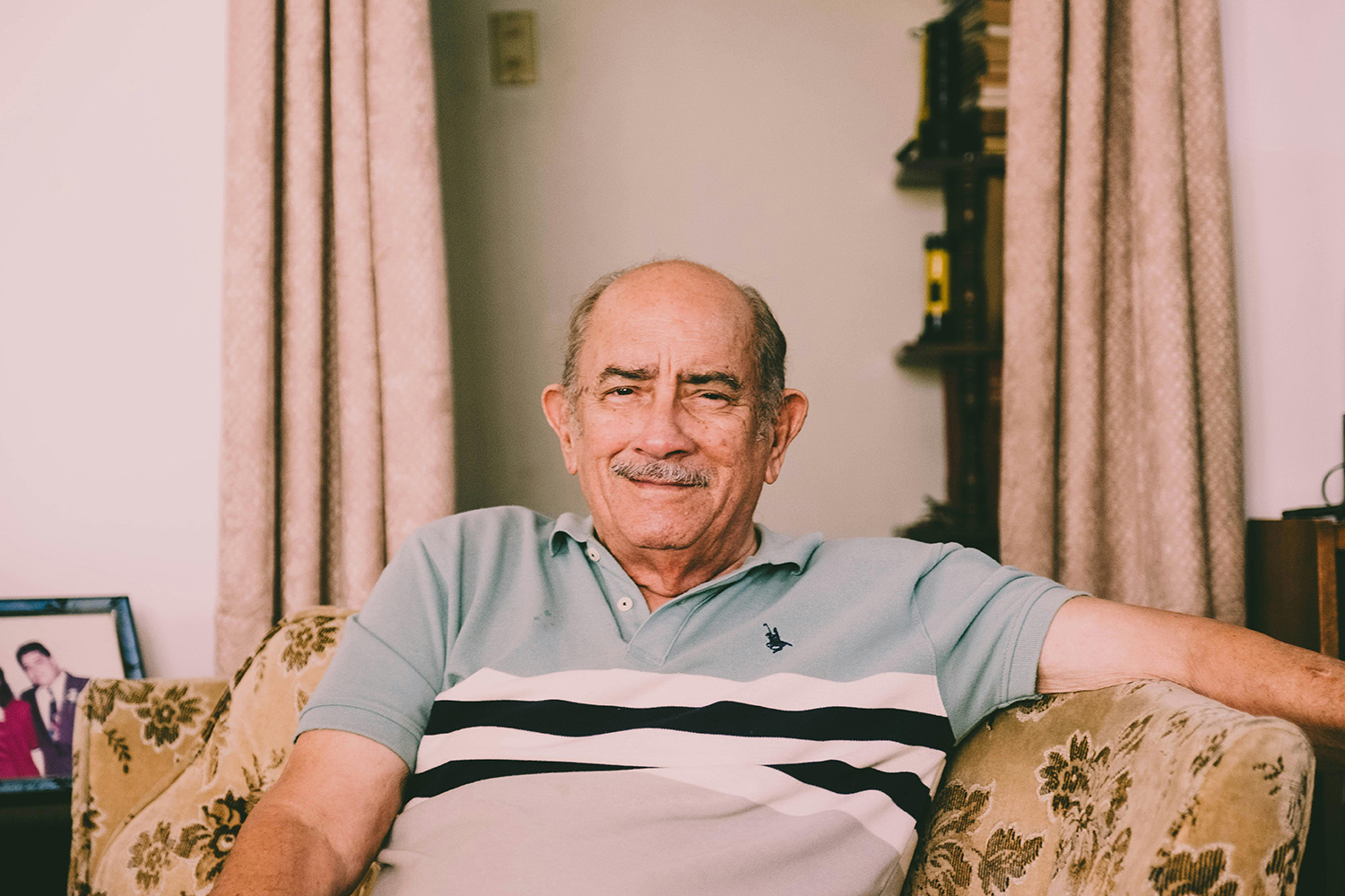 A portrait-style photograph of an elderly man sitting in what appears to be a living room. He smiles timidly at the camera. This image represents the accessibility and housing challenges faced by persons with disabilities.
