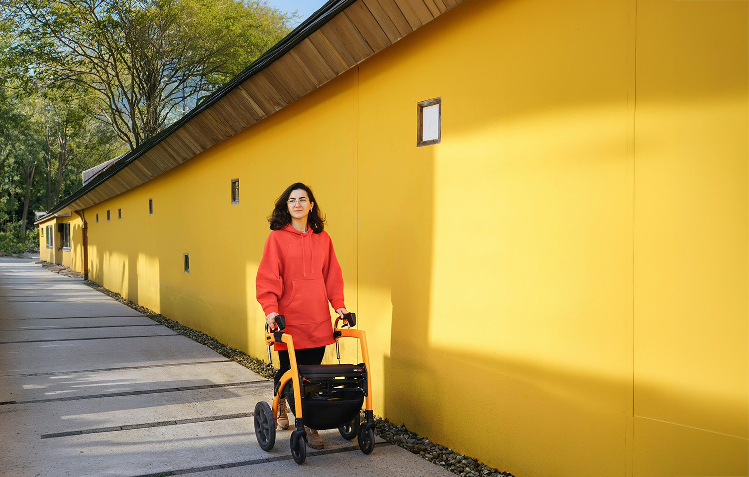 A young person in a red sweatshirt walks outdoors using a walker for mobility support. They look off-camera pensively, with wind blowing through their hair.