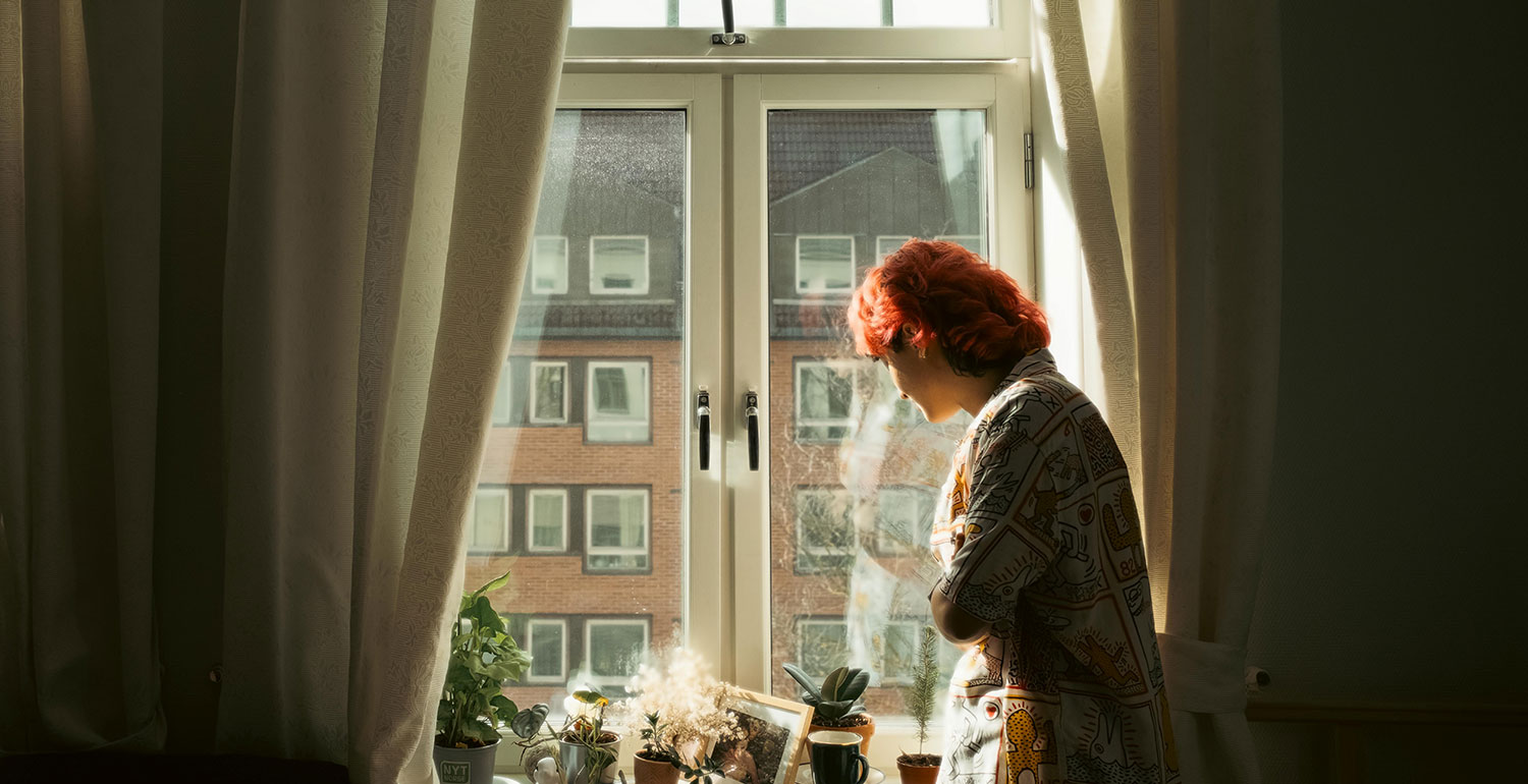 A person stands with their back to the camera, looking out the window toward the street. The tones and mood are pensive. The image represents Adya, the person in the case story.
