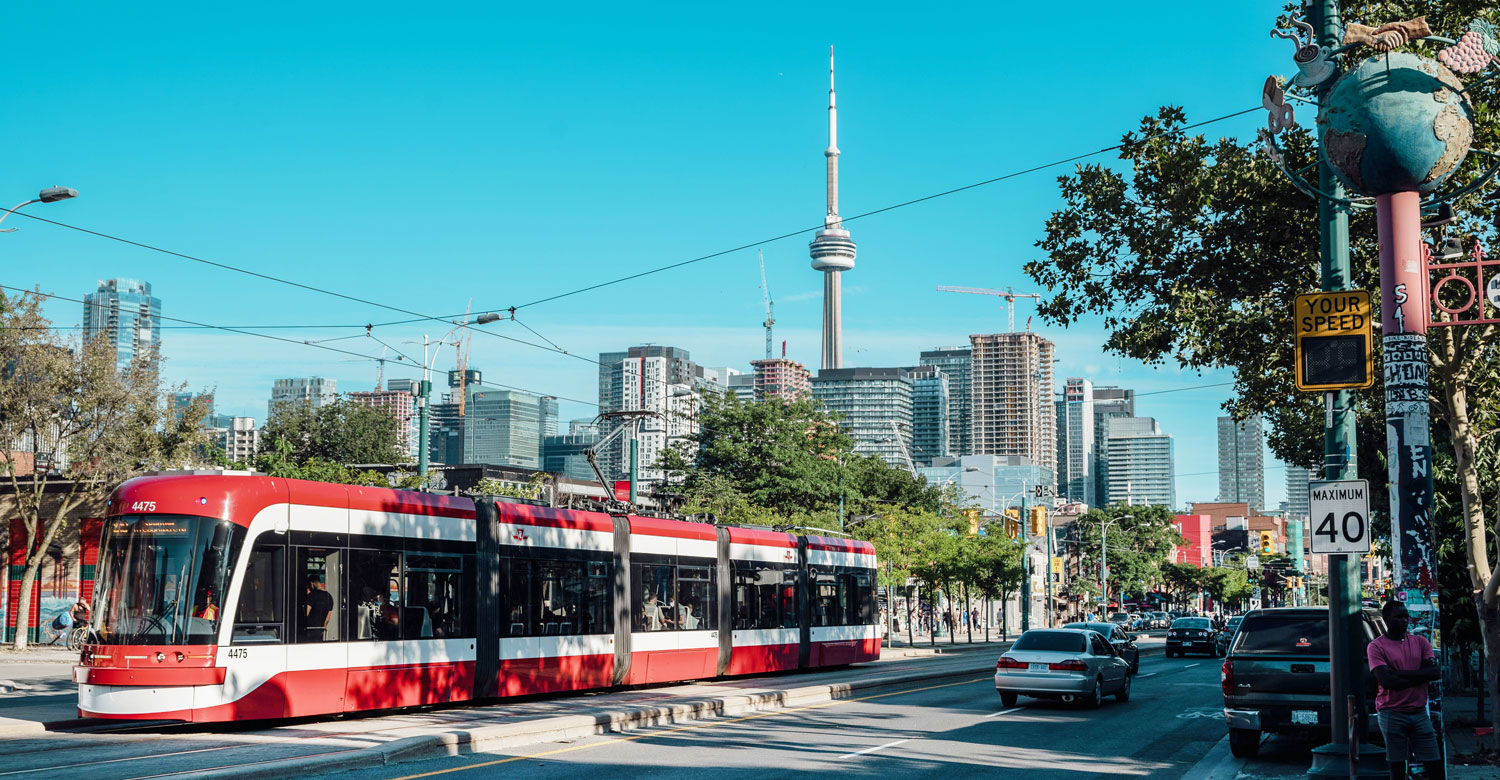 A red and white TTC streetcar is traveling down a street in Toronto. The streetcar is passing by a tall building and a street sign. There are also cars on the street, including one in front of the train and another behind it.