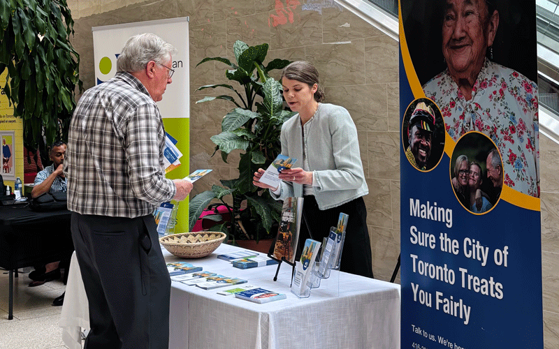An Ombudsman Toronto staff member speaking to a member of the public, a banner next to her reads "Making sure the City of Toronto treats you fairly"