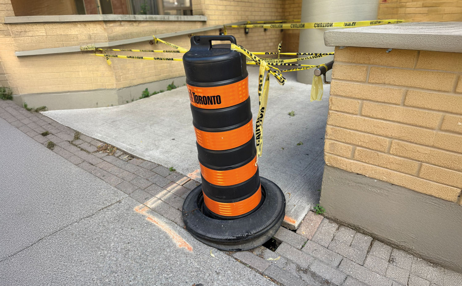 A barricade is set up in front of a building, with a large orange and black barrel and a cone placed in front of it. The barricade is marked with yellow tape.