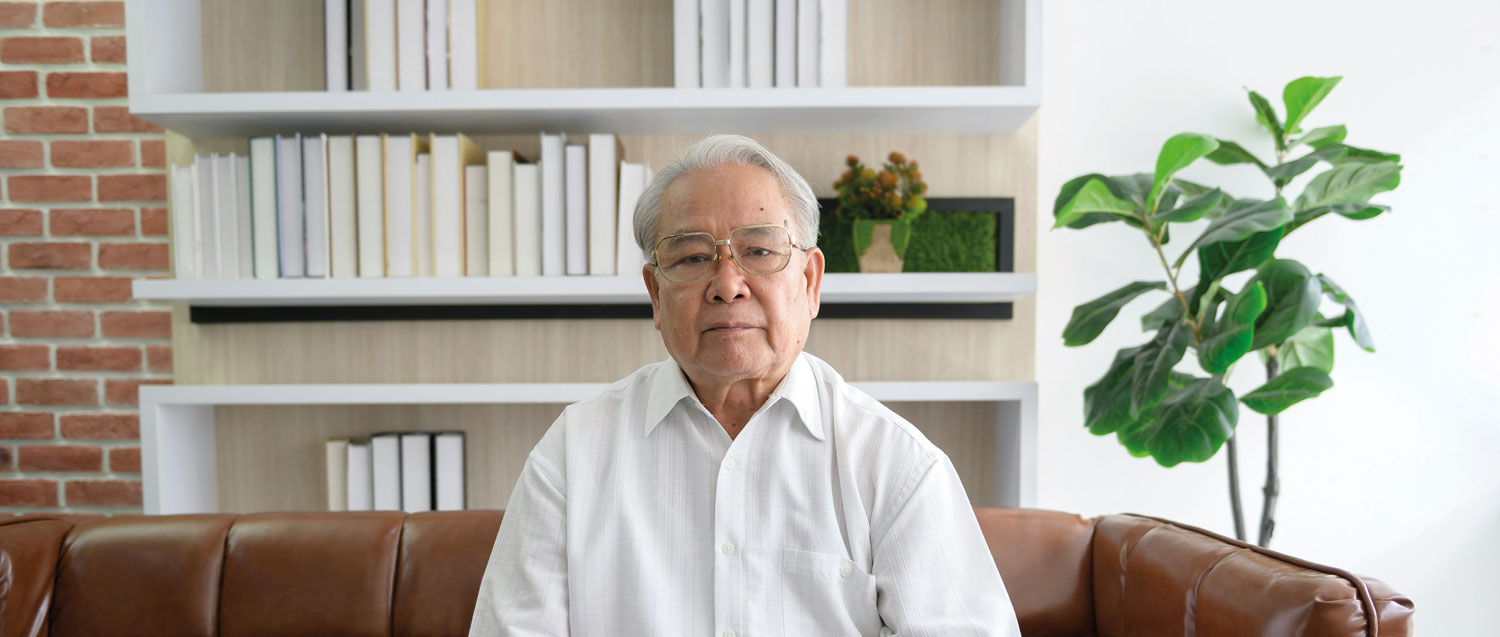 An older man is sitting on a brown couch, wearing a white shirt and glasses.