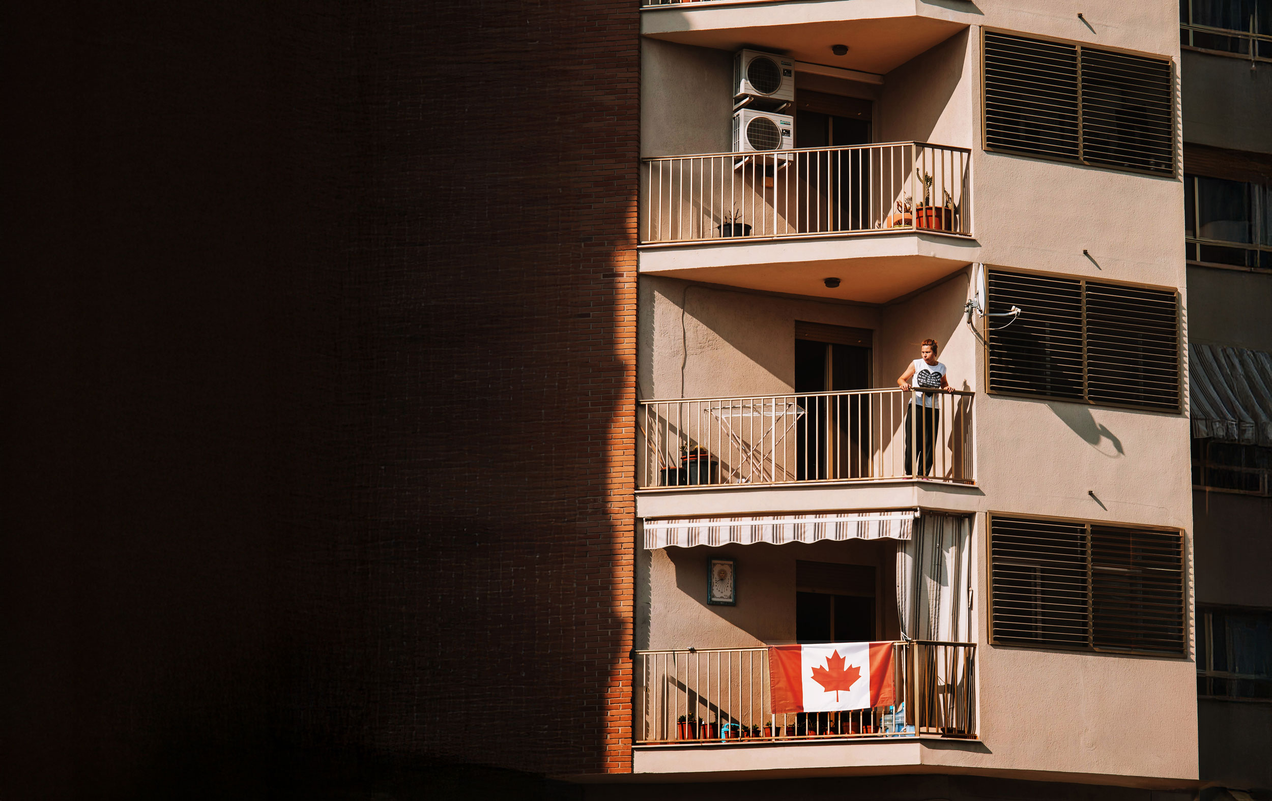 Person stands on a balcony outside of their apartment, looking down at the street. A balcony below shows a Canadian flag hanging off the banister.